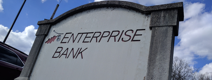 The words "Enterprise Bank" are engraved in a stone marker. The blue sky is visible in the background.